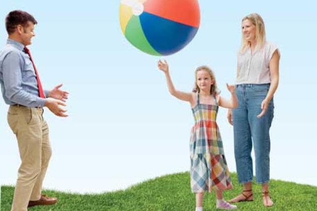 A young girl tosses a large inflatable beach ball outside with a man and a woman. The adults are smiling. The girl concentrates on throwing the ball to the man.
