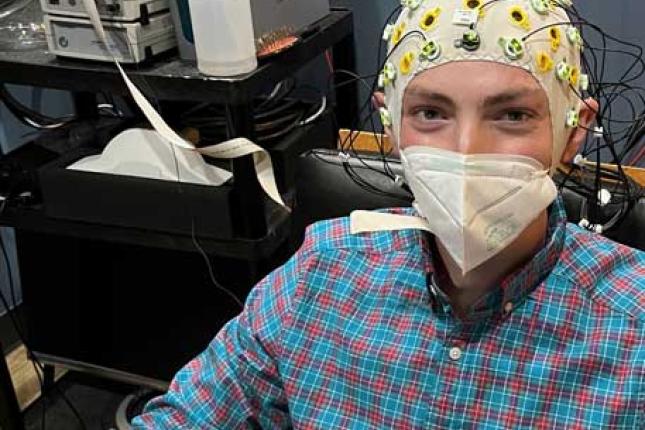 A young man sits in chair in a clinical room. He wears a face mask and a snug-fitting EEG cap fitted with dozens of electrodes. 