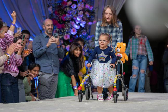 Penny, a child in colorful clothing, walks down a runway with assistance while lots of people in a crowd smile and watch.
