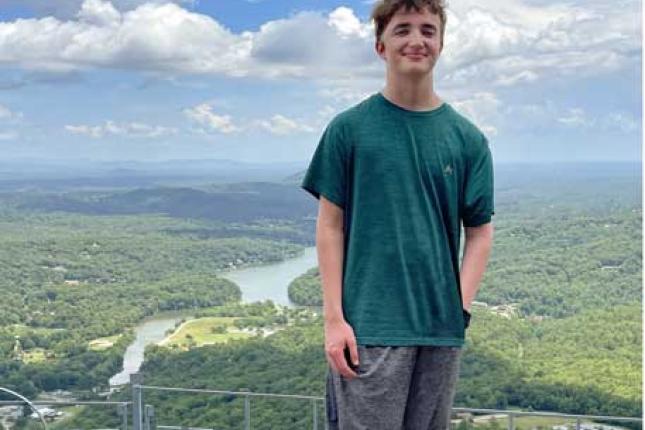 A young man smiles while standing atop a hill or summit. Behind him is a beautiful landscape, with trees, mountains and a river. 