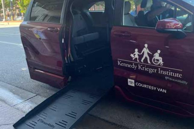 A wheel-chair accessible Toyota Sienna is parked at the curb with the ramp lowered. The adaptive van is burgundy and has Kennedy Krieger Institute's logo on the front passenger door. 