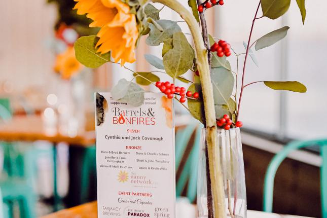 A vase with flowers and plants in it, next to a card that reads Barrels & Bonfires.