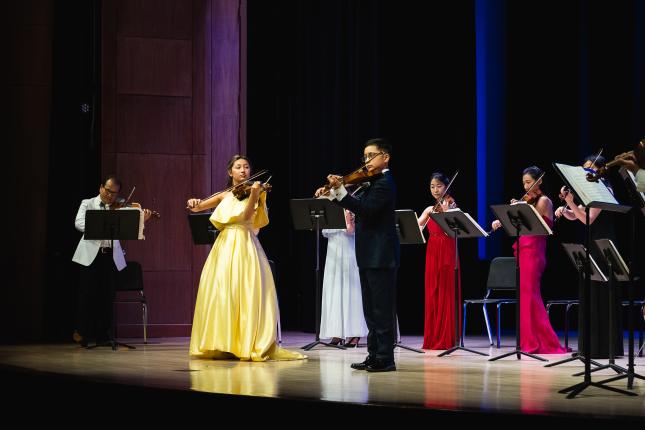 Siblings Maggie and Matthew, wearing a yellow dress and black suit respectively, stand on a stage playing violins with an ensemble in the background.