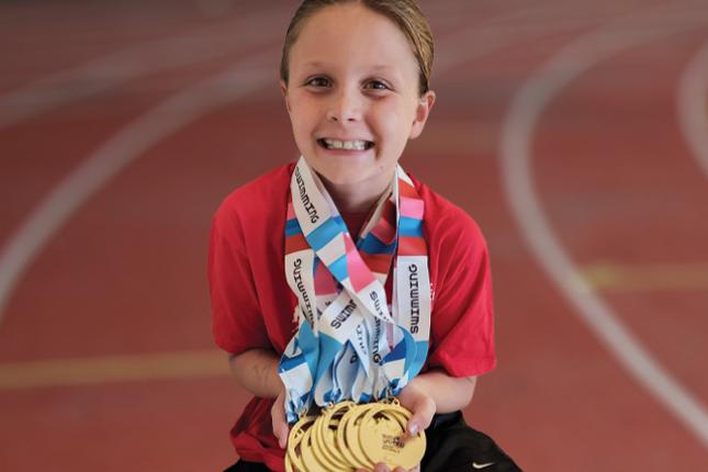 Penelope, smiling, wears nine medals around her neck, all of them gold, and with red, white and blue ribbons. Some of the ribbons can be seen to say “SWIMMING” on them.