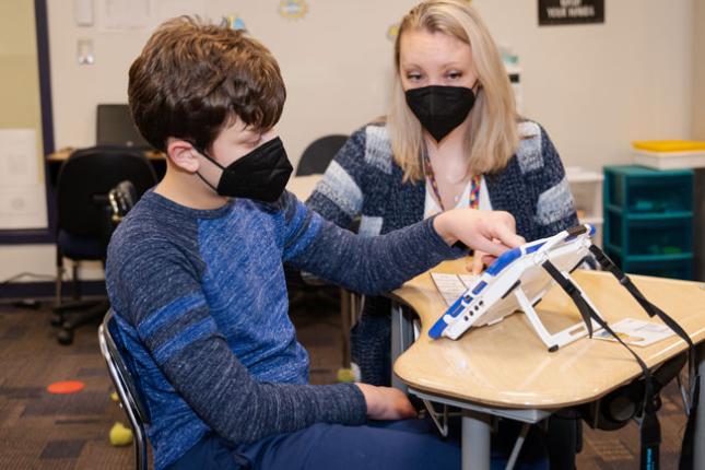 Jack sits at a student desk in a classroom with his teacher and uses a tablet-like communication device.