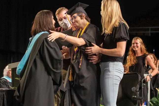 Hailey stands on the stage at her high school graduation and reaches out to hug her school principal. Hailey is smiling and wears a black graduation cap and gown, a medal, and purple and gold academic cords draped over her shoulders. Her school principal is also smiling and wears a black academic gown with a hood lined with light blue velvet. Two therapists stand to either side of Hailey, ready to help her if she needs assistance walking or standing. Behind the group is Hailey’s sister, who is smiling and h