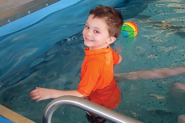 Tripp smiles during aquatic therapy. An inflatable ball floats in the pool behind him.