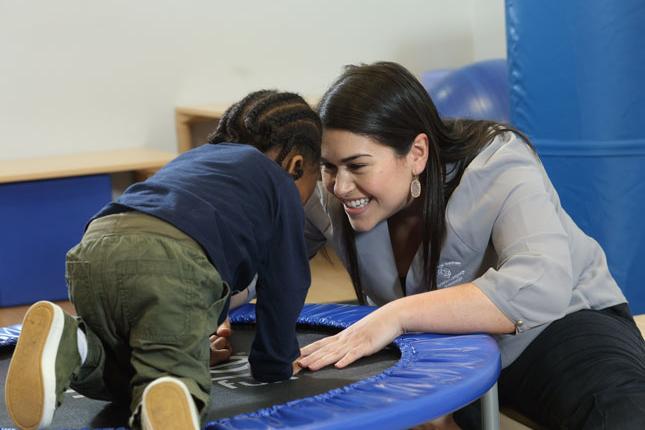 A child with autism plays on a small trampoline, while a woman at one end of the trampoline smiles at him. 