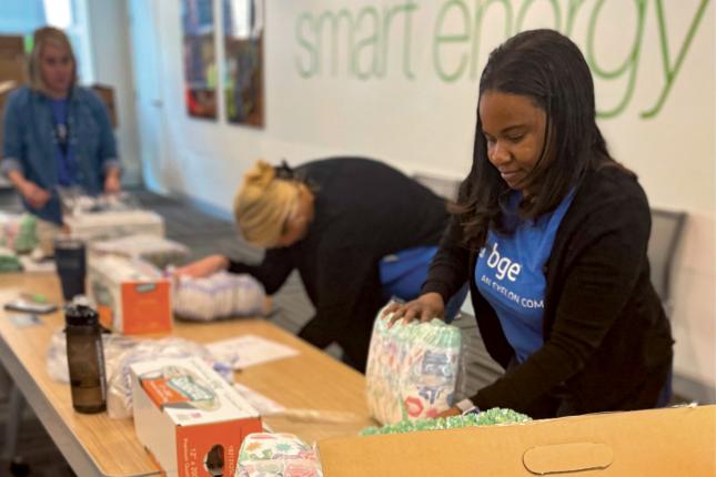 Two women work at a table assembling diapers into care packages. Each woman is wearing a blue T-shirt. One of the T-shirts can be seen to read “BGE, an Exelon Company.”