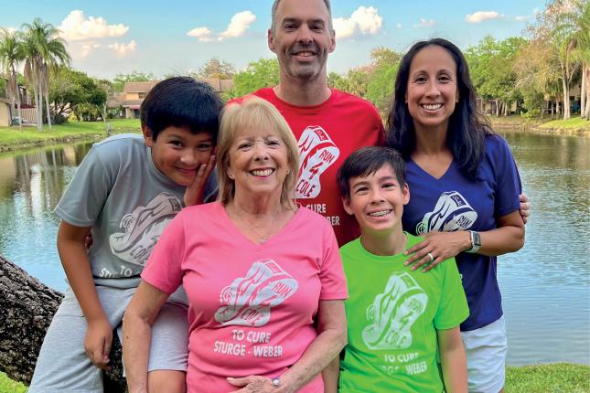 Portrait photo of a family outside, with a pond and palm trees in the background. The family members are Mom, Dad, Grandma and two boys. Each person wears a T-shirt with “Run 4 Cole to cure Sturge-Weber” on it. Each T-shirt is a different color.