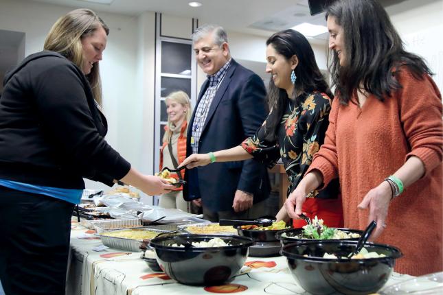 Four people stand on one side of a table, serving food. On the other side, a woman receives a plate of food.