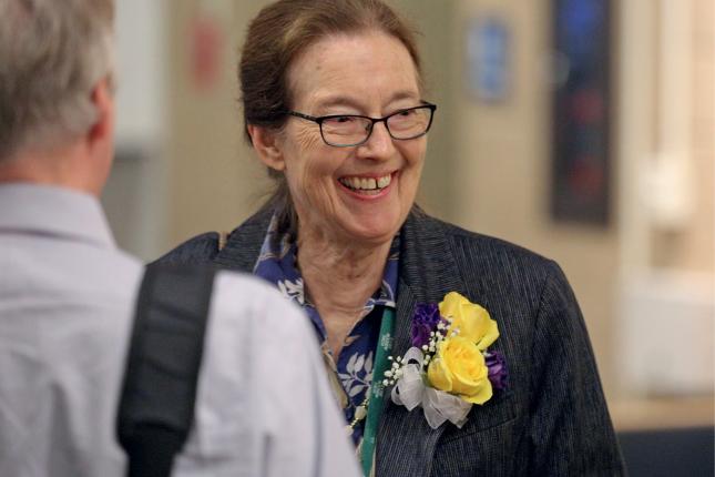 A woman smiles and looks off to the side. She is wearing a corsage of yellow roses and purple flowers, with a white bow. In the foreground is a man’s blurry back. They are standing in a room or hallway.