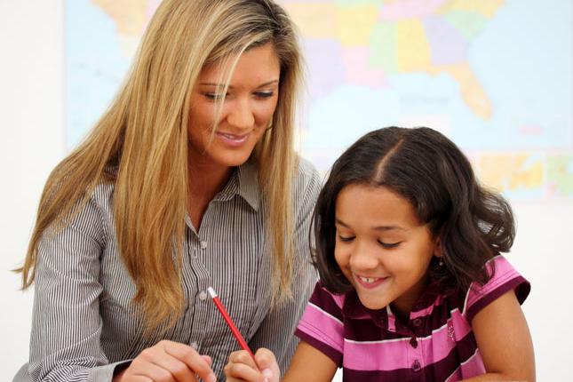 A woman teacher sits next to a young girl while she writes. 