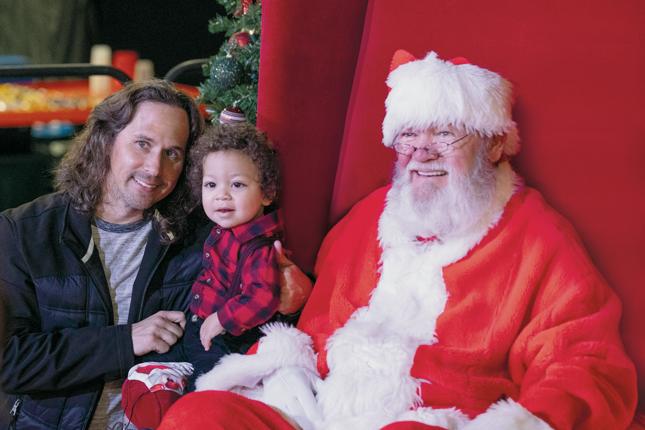 A father holds his toddler while kneeling next to Santa Claus at Festival of Trees.
