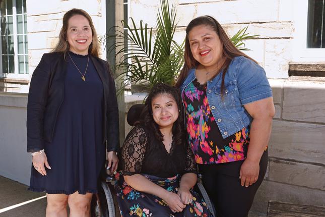 A teenaged girl in an elegant outfit sits in a wheelchair between two women. She leans toward the woman on the right, who leans toward the girl, as they are mother and daughter. All three are smiling.