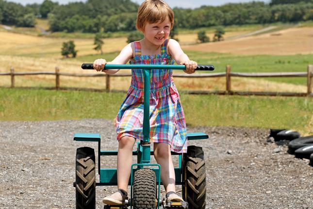 A smiling little girl with short blonde hair rides a tricycle in a rural setting.