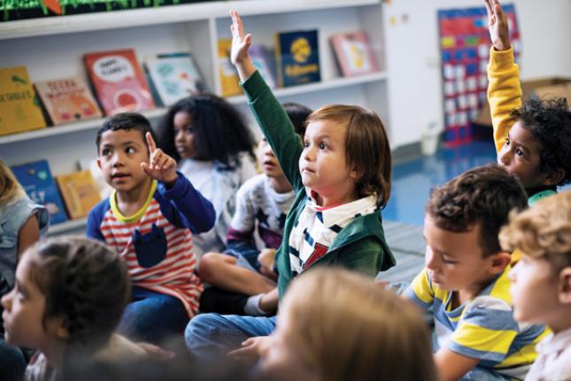 Three kids sitting on a floor in a classroom raise their hands. Several students sitting around them are not raising their hands.