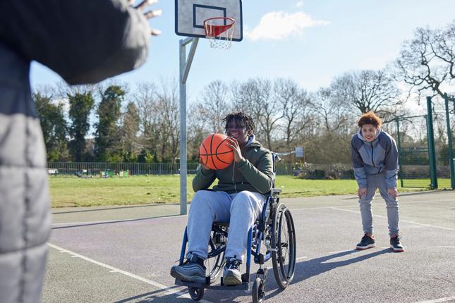 A teenager using a wheelchair plays basketball at an outdoor court with two other teenagers.