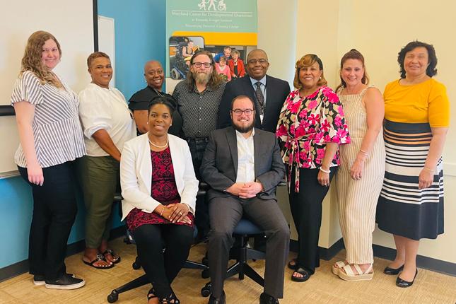 A group photo from a Faith Community meeting. A woman and man sit in the front with a group of four people standing behind them. The top of a green and yellow MCDD banner can be seen in the very back.