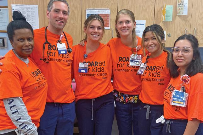 Six smiling people pose for the camera wearing bright orange shirts that say “I ROARed for Kids at Kennedy Krieger 2022.” Most of the people have badges on; some of the badges say “RN” on them. One of the people has a stethoscope around their neck.