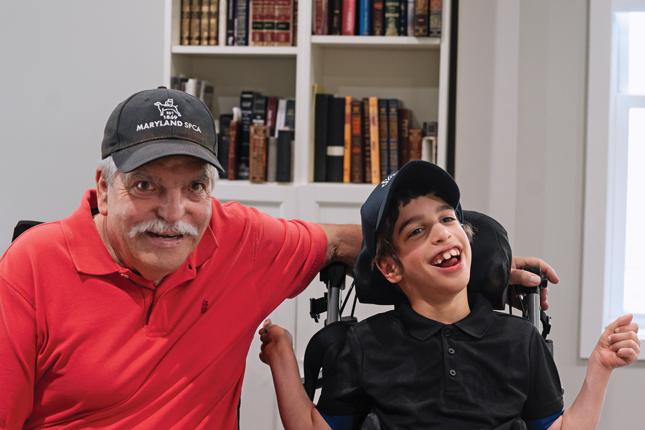 A man wearing a baseball hat that says “Maryland SPCA” on it sits beside a young boy who is also wearing a baseball cap. The boy is sitting in a wheelchair. The man’s left arm rests on the back of the wheelchair, behind the wheelchair’s headrest. Both the man and the boy are smiling. They are sitting indoors, in front of a bookshelf.