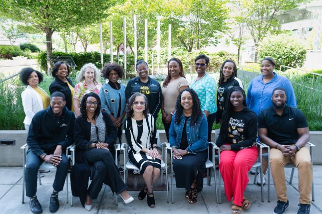 A group of 15 people pose in a garden-like setting. Six of them are sitting in chairs, while the other nine stand behind them. All are smiling, and most are people of color.