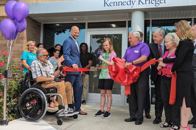 A group of at least 16 people of different ages gathers outside a glassed-in entrance with “Kennedy Krieger” above the doors. The group is holding a long, wide red ribbon with a giant bow in the center. A young person wearing a shirt that says “HOPE” is cutting the ribbon with giant scissors. Holding one end of the ribbon is a man sitting in a wheelchair. Some people are wearing suits, while others are wearing more casual clothing.