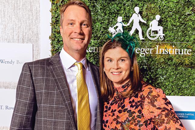 A husband and wife wearing fun formal wear pose for the camera, smiling. Behind them is a clear sign with Kennedy Krieger Institute’s logo on it. The sign hangs in front of a leaf-covered wall.