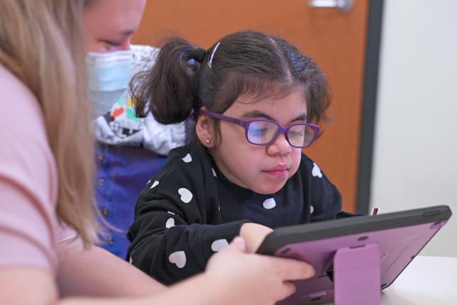 A young girl, assisted by a therapist, uses a tablet-like device that helps her communicate.