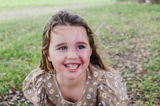 Rainey leans forward and smiles at the camera while sitting on the ground outside. 