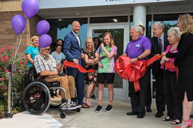 A group of people gathering around a young girl as she cuts a red ribbon to open the International Center for Spinal Cord Injury's White Marsh location.