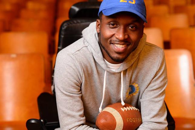 Van Brooks sits in a wheelchair while a football rests in his lap. He is wearing a blue hat with the number 25 in yellow.