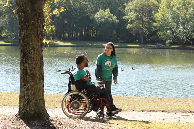 A man and woman talk on a pathway that winds around a pond. The man is sitting in a wheelchair, while the woman is standing to his left looking at him.