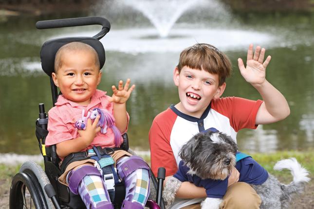 Posed photo of a young boy sitting in a wheelchair and a slightly older boy holding a small dog. Both boys are smiling and waving.