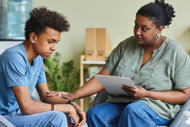 A woman holding a clipboard and with a concerned expression on her face places one of her hands on the arm of a teenage boy.