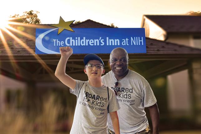 Posed photo of two smiling men in front of a building with a sign that says “The Children’s Inn at NIH.”