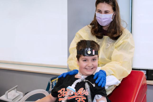 Posed photo of a smiling boy with a woman behind him. The woman is wearing a yellow medical gown and a face mask and has her hands on the boy’s shoulders. Her squinting eyes indicate she is smiling under her face mask. The boy is smiling and wearing a shirt that says “Orioles” on it. He has clear plastic tubes attached to his neck for breathing purposes and is wearing a head lamp-like device. Two electrodes marked “Leo” are attached to his left arm.