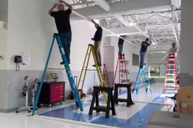 Five adults stand atop colorful ladders in an open interior space while adjusting ceiling apparatus.