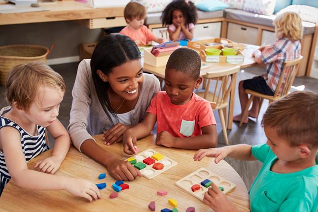 A teacher smiles as she helps three pre-school students with an activity at their desk.