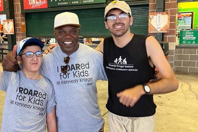 Three men stand on the concourse of a stadium smiling. The man to the left has his right fist raised in the air.