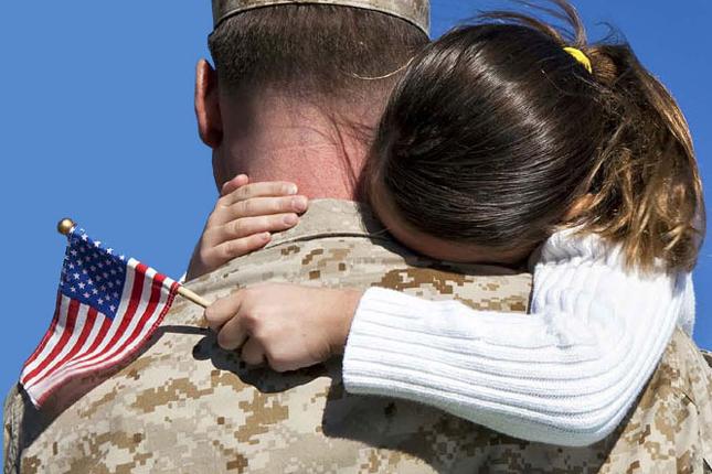 A soldier carries his young daughter, who is wrapping her arms around his soldiers while holding a small United States flag.