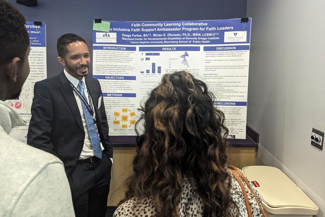 A man stands in front of a research poster and speaks to two people. The other two individuals have their backs to the camera.