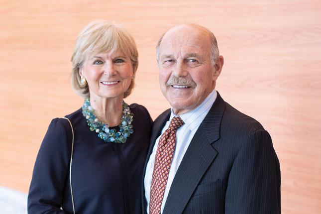 A woman and a man smile for the camera. The woman is wearing a black dress and necklace of blue and green glass beads. The man is wearing a black suit with a red tie.