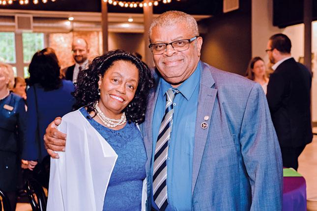 A woman and a man smile for the camera. The man is taller and to the right, and he has his right hand around the woman’s right shoulder. The woman is leaning into the man’s right shoulder. The man is wearing a blue suit, tie and glasses, and the woman is wearing a blue dress and pearl necklace.