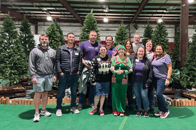 A group of 11 people of various ages, many wearing purple and one wearing a green Christmas elf costume, pose in front of a group of artificial trees inside a large indoor space, which looks like it might be a convention hall.