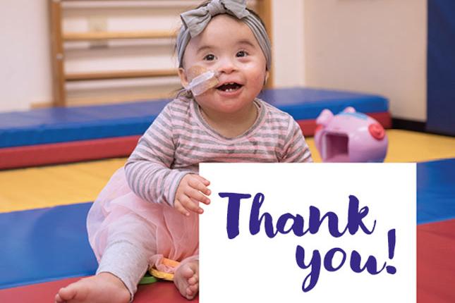 A toddler sits on the floor, with her left hand holding a white sign that says Thank you! in purple font.