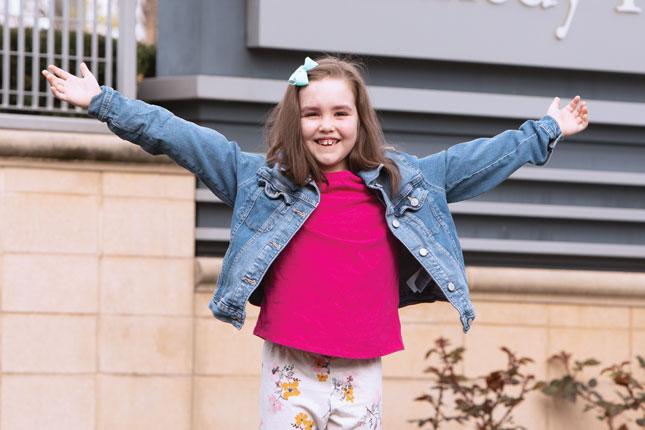 A young girl smiles with her arms outstretched. She has faint port wine-colored birthmarks across her forehead. A sign with the words “Kennedy Krieger” is in the background.
