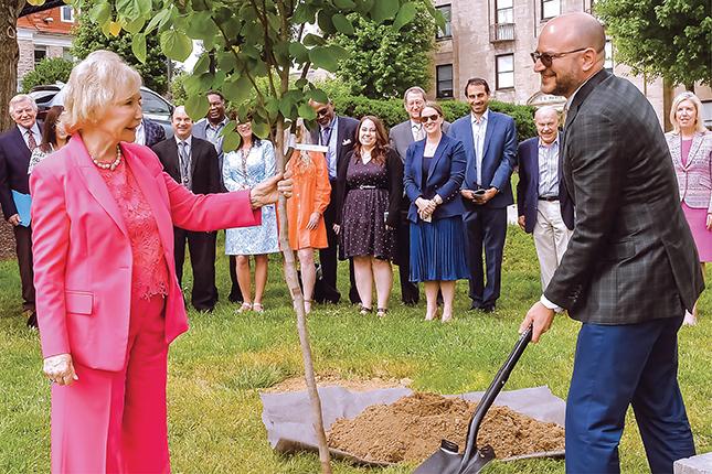 A woman and a man in business attire plant a tree, with a group of people standing behind them.