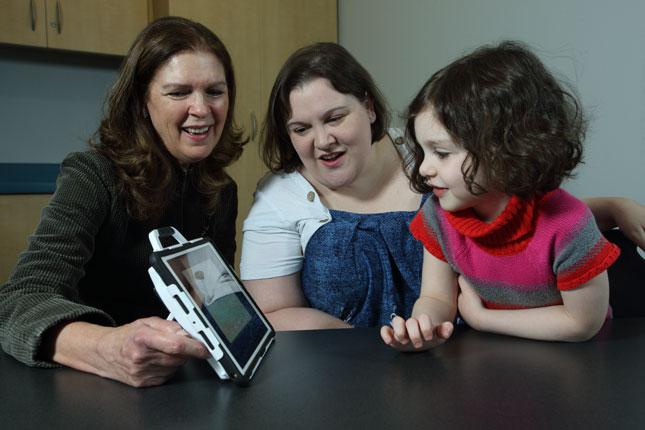 Una médica sonríe mientras enseña a una niña un juego en una tableta. La niña está en el regazo de su madre, y la madre y la médica están sentadas ante una mesa.