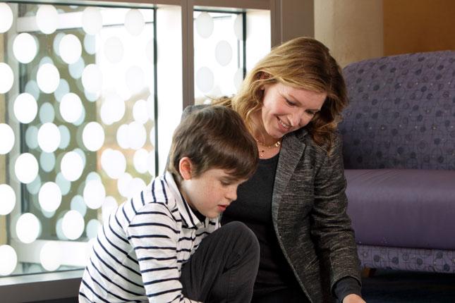 A boy with a muscular dystrophy sits on the floor next to a woman while reading a book.